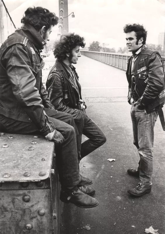 Black and white photograph of three young men in leather jackets, jeans and boots on Chelsea Bridge, two sitting on a riveted girder while the third stands facing them.