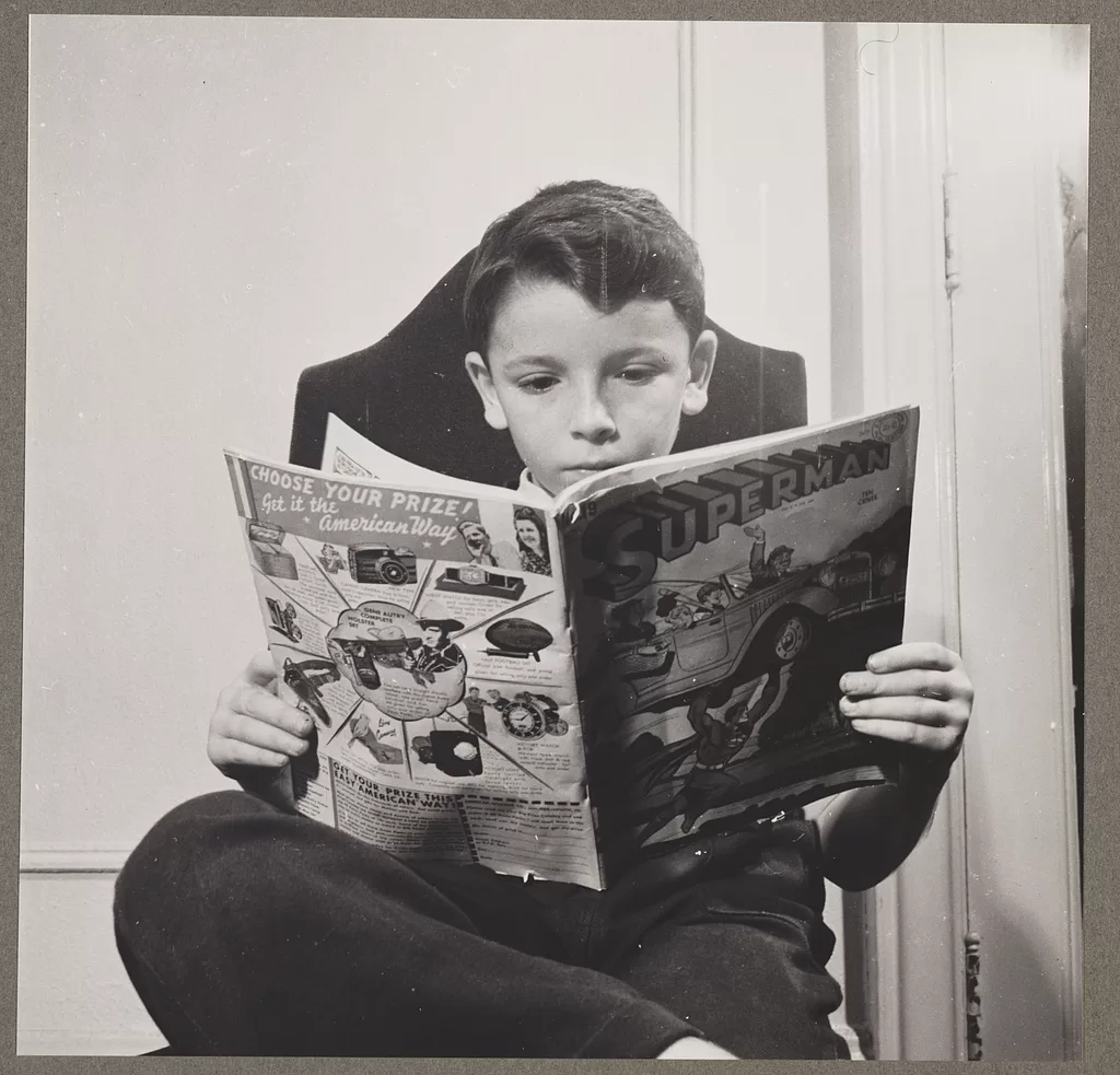 Black and white photograph of a boy sitting cross-legged on a chair, absorbed in a Superman comic, its back cover advertising prizes to "Get it the American Way".